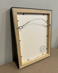 The back of the framed drawing of Shadow Bloom by Kris Horton showing the wooden frame and the wire hanger.
