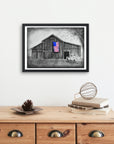 Framed artwork by Kris Horton of a barn with an American flag on a white wall above a wooden cabinet.