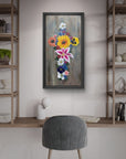 Framed floral painting of a cross made of flowers by Kris Horton, on a wall above a desk with shelves and a chair.
