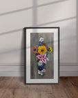 Black framed painting by Kris Horton of a cross made of colorful flowers with a gray background leaning against a wall.