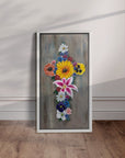 White framed painting by Kris Horton of a cross made up of colorful flowers leaning against a wall.