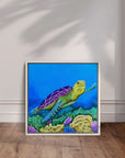 White framed painting by Kris Horton of Sea of Colors a turtle swimming in blue water above coral, displayed leaning against wall.
