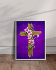 White framed artwork of a wooden cross with pink flowers on a purple background, displayed leaning on a wall.