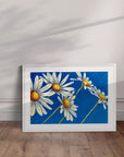 White framed Daisy Days artwork by Kris Horton of daisies on a blue background in a room with wooden floor and white walls.
