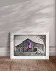 White framed painting by Kris Horton of My America a monochromatic rustic barn and old chevy truck with an American flag in color leaning against a wall.