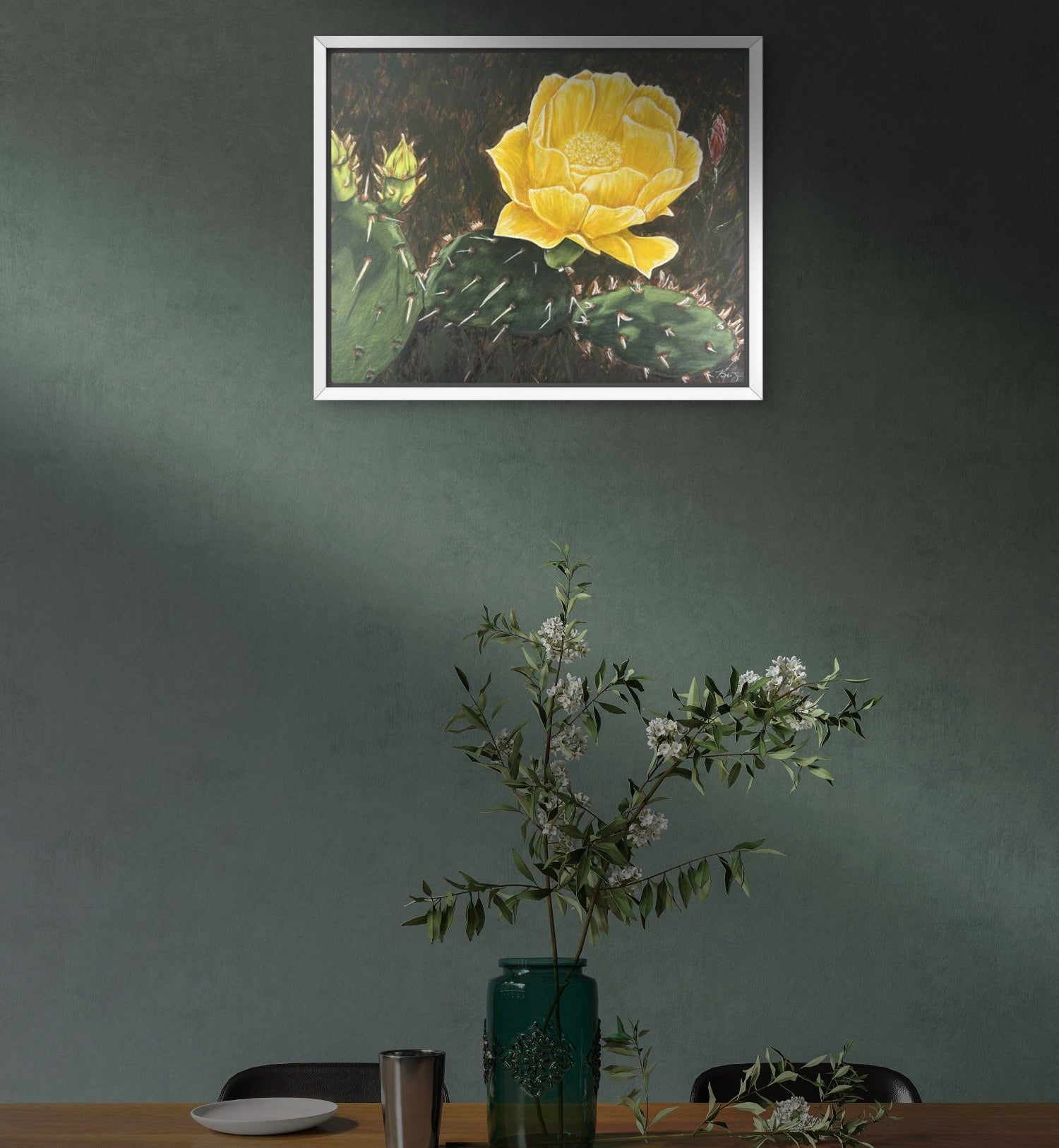 Framed artwork of a yellow flower on a wall above a table with a vase and plants, painting by Kris Horton of a cactus bloom