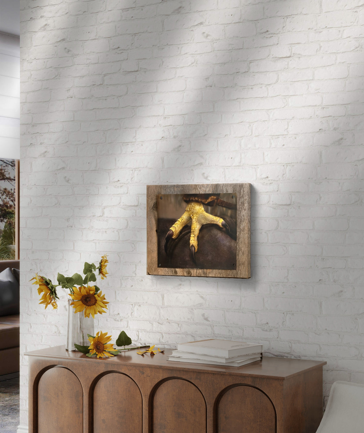 Framed painting of Talon a close up of a red tail hawks foot by Kris Horton, on a white brick wall above a wooden console table with sunflowers.