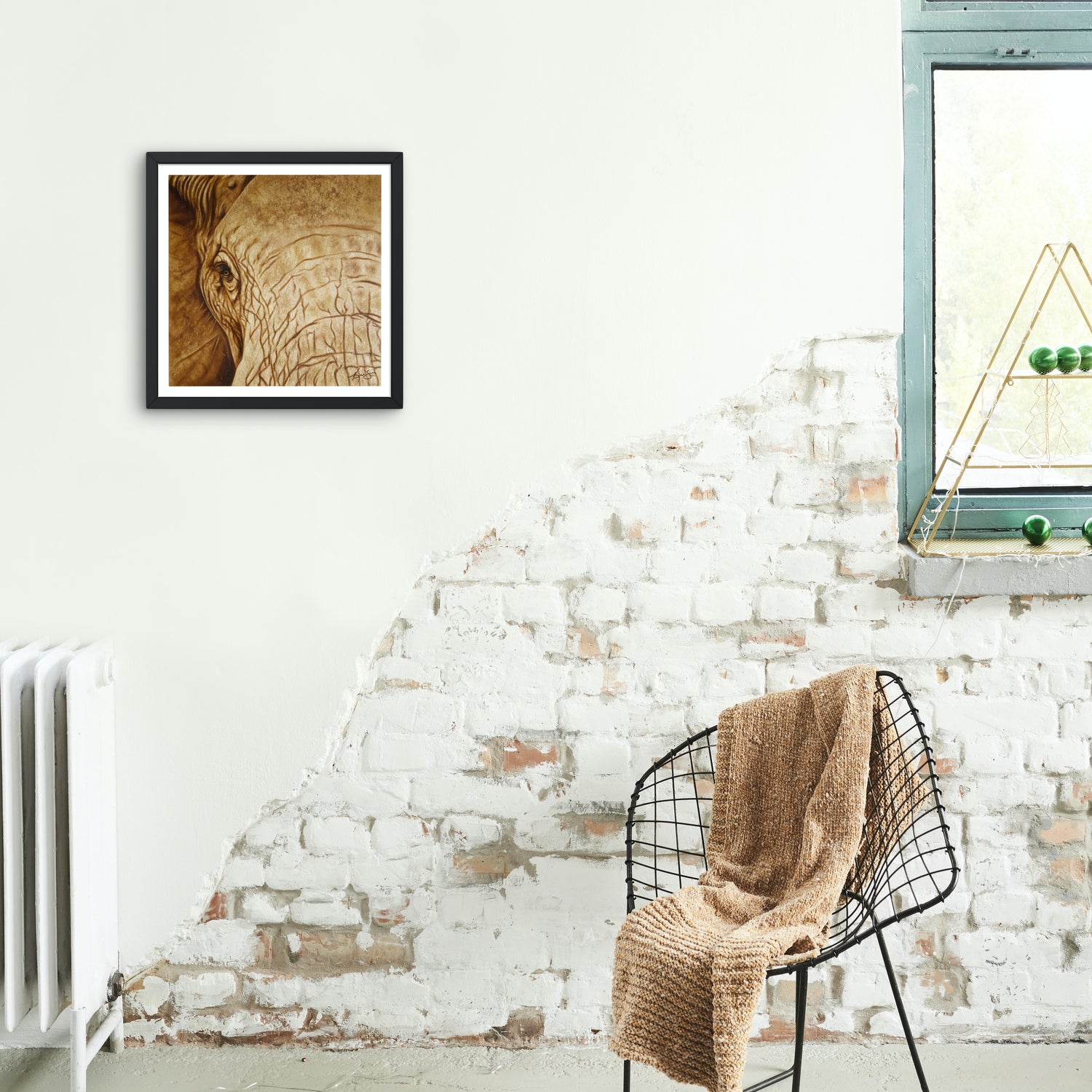 Framed wall art by Kris Horton of an elephants face in sepia hanging on a white brick wall.