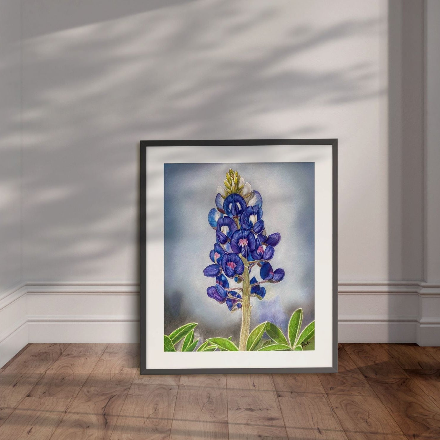 Framed artwork of a bluebonnet flower painting by Kris Horton leaning against a wall.