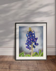Framed artwork of a bluebonnet flower painting by Kris Horton leaning against a wall.