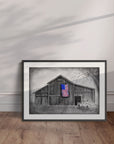 Framed artwork by Kris Horton of a barn with an American flag leaning against a wall.