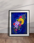 Framed artwork of colorful flowers on a blue background displayed leaning against a wall.