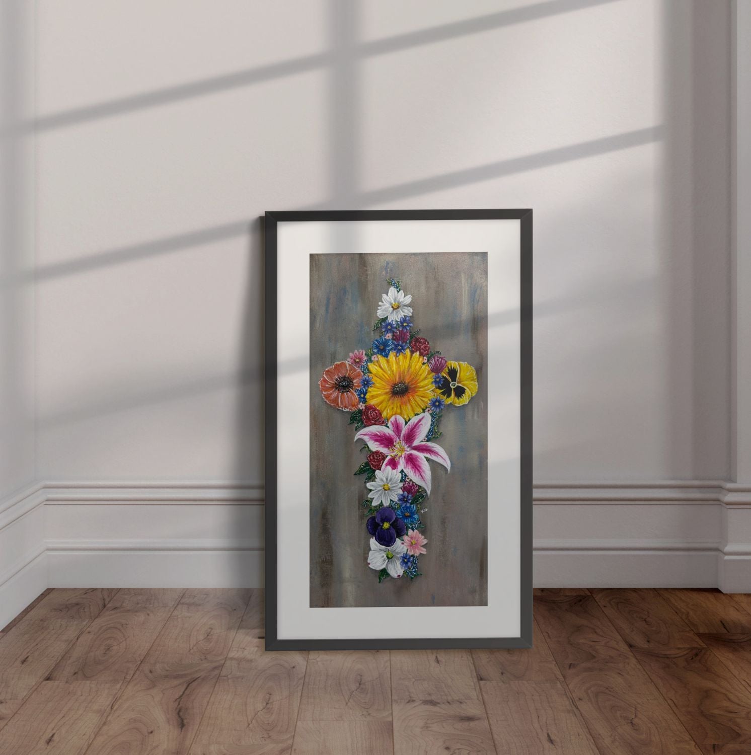 Black framed painting of a cross made of colorful flowers with a gray background leaning against a wall.
