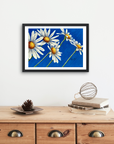 Framed artwork by Kris Horton of Daisy days which is daisies on a blue background above a wooden cabinet.
