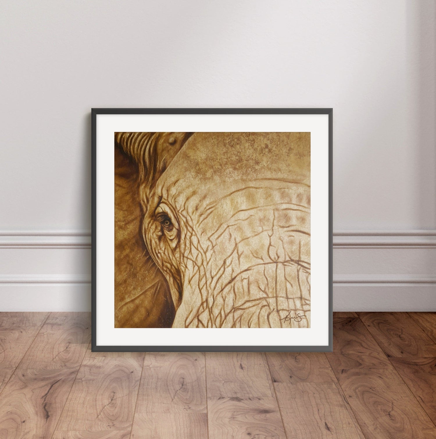 Black framed artwork by Kris Horton of an elephant's face leaning against a wall on a wooden floor.
