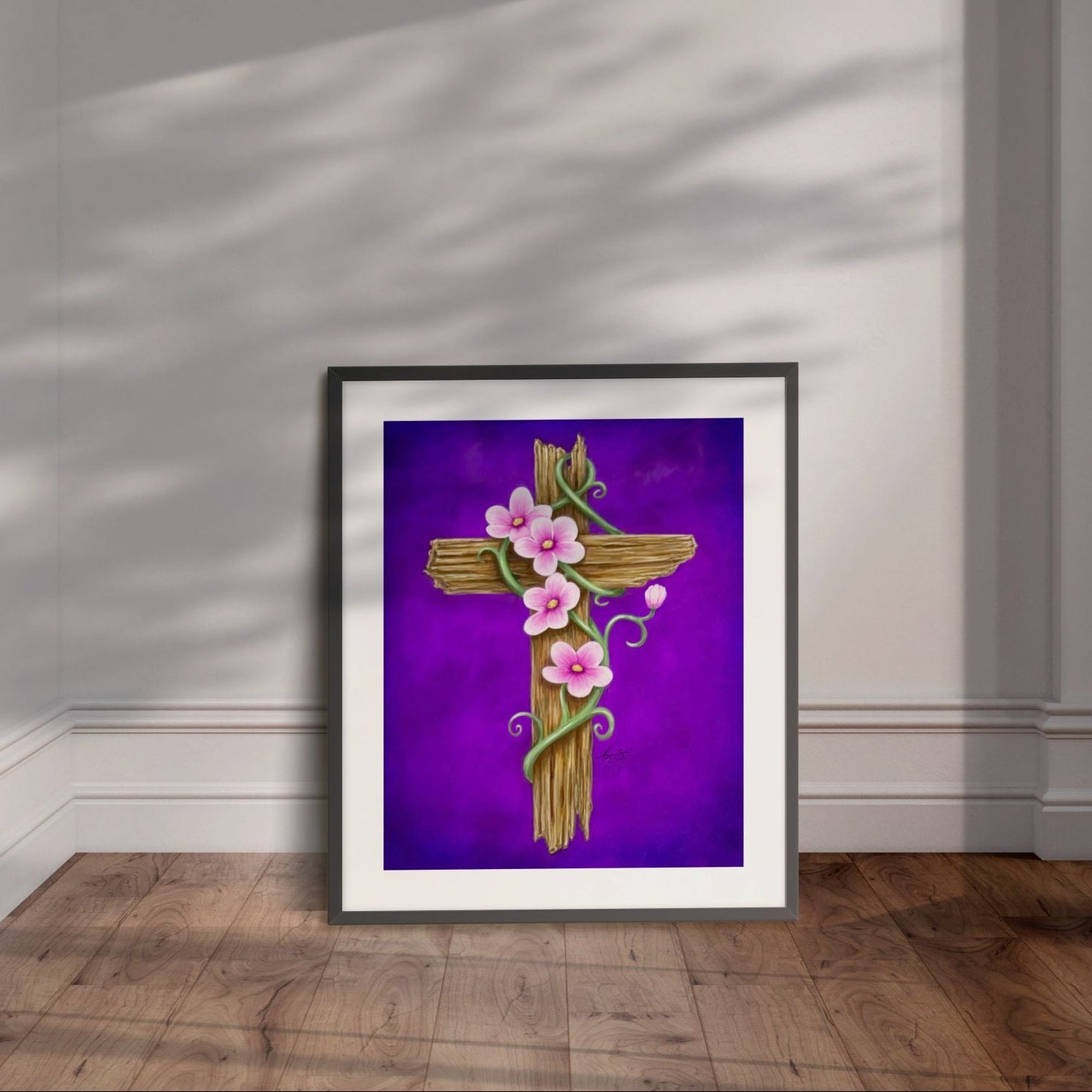 Framed artwork by Kris Horton of a wooden cross with pink flowers on a purple background, displayed leaning on a wall.