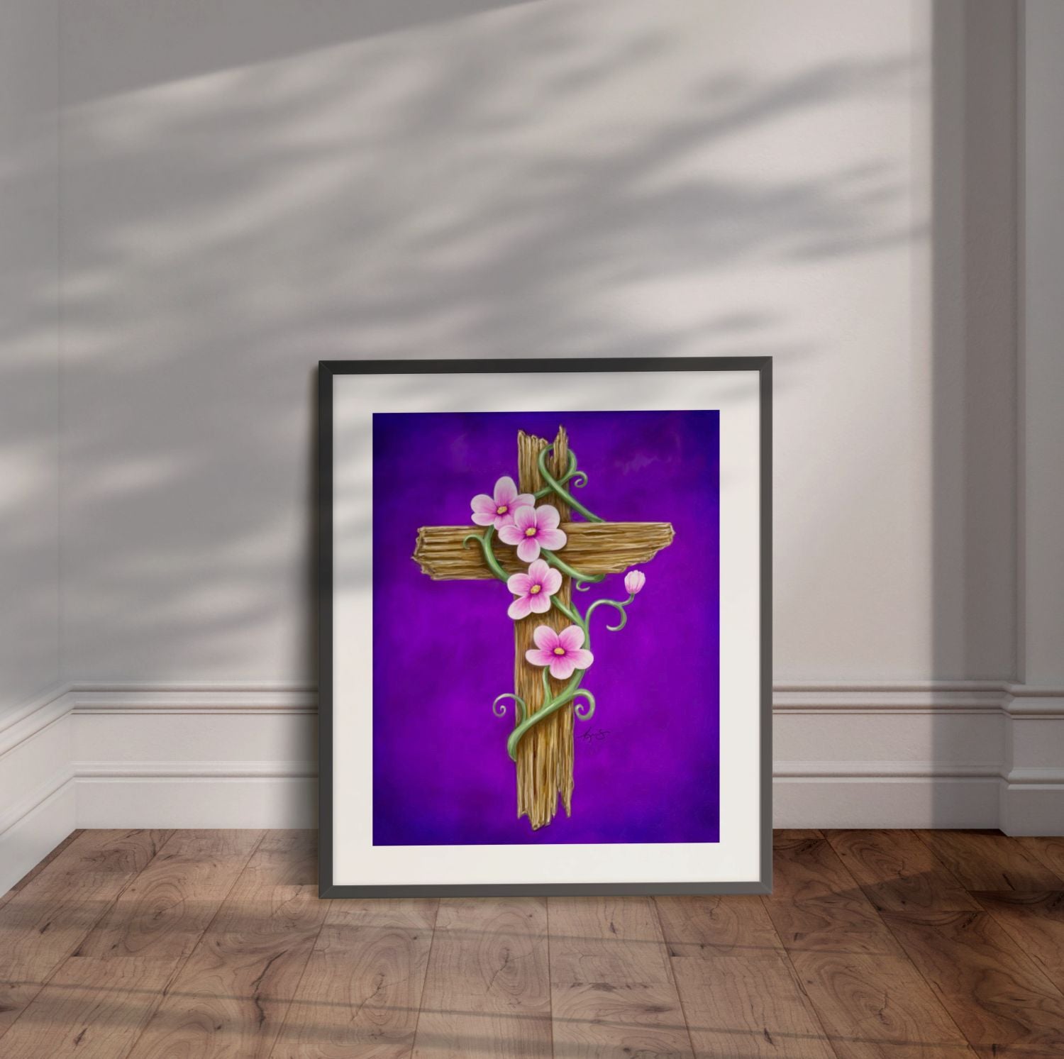 Framed artwork of a wooden cross with pink flowers on a purple background, displayed leaning on a wall.