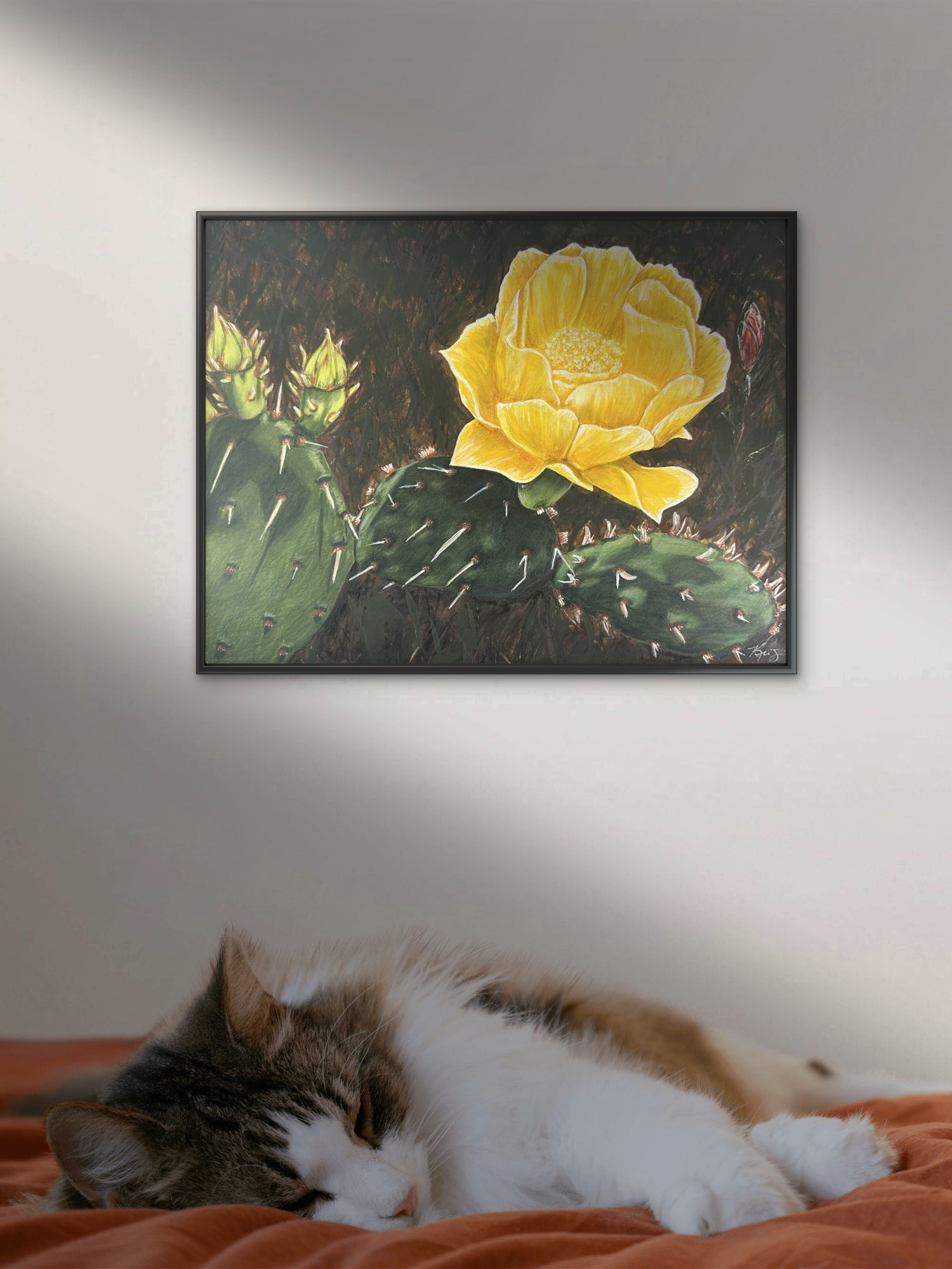 Framed artwork of a yellow cactus flower on a wall above a cat lying on a bed painting by Kris Horton
