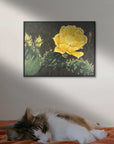 Framed artwork of a yellow cactus flower on a wall above a cat lying on a bed painting by Kris Horton