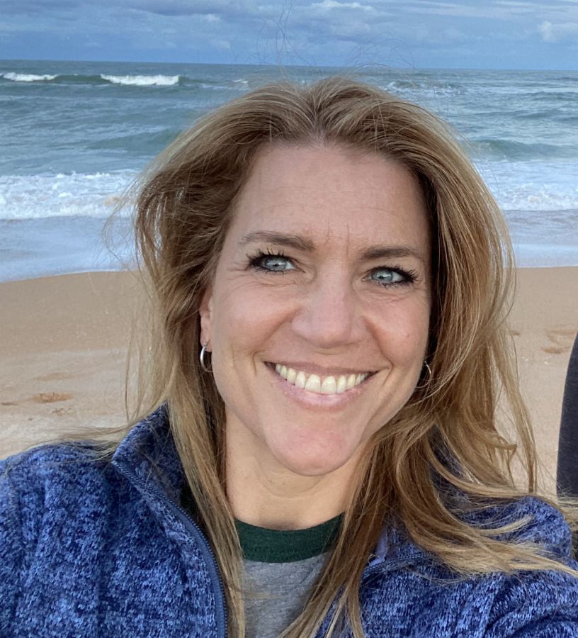 Kris Horton, the artist, smiling on a beach with ocean waves in the background