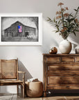 Framed monochromatic painting by Kris Horton of a barn and truck with an American flag in color hanging over a chair and wooden sideboard.