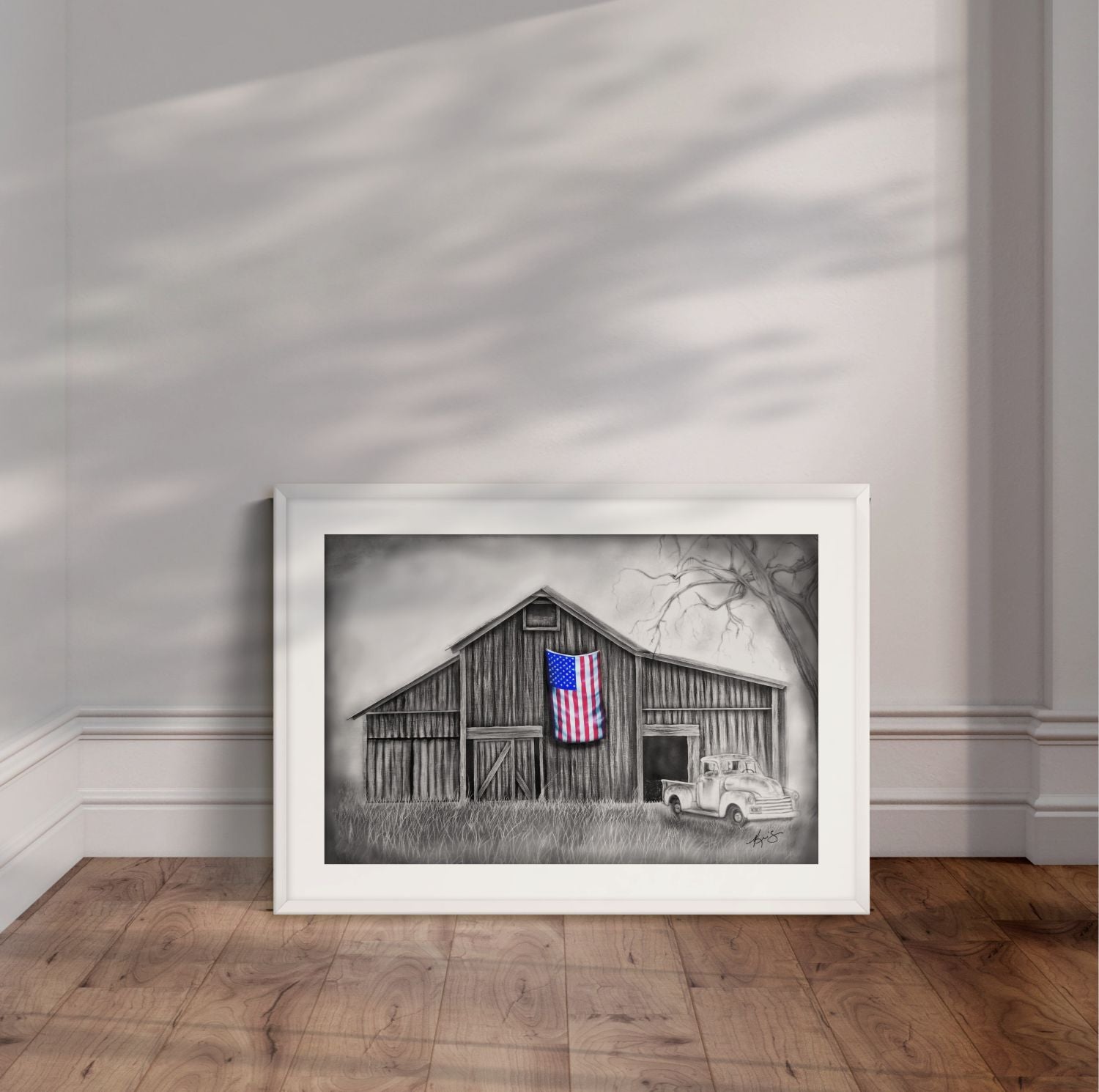 White framed painting by Kris Horton of My America a monochromatic rustic barn and old chevy truck with an American flag in color leaning against a wall.