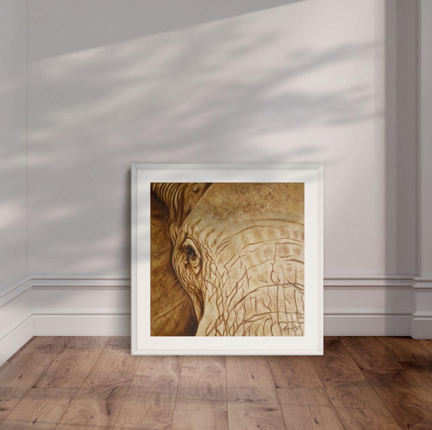 White framed artwork by Kris Horton of a close up of an elephants face leaning against a wall.